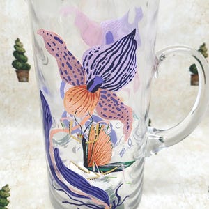 Puede incluir: Jarra de vidrio transparente con un dise&ntilde;o floral en tonos rosa, morado y naranja. La jarra tiene un asa curva y un pico vertedor. El dise&ntilde;o floral incluye una mariposa.