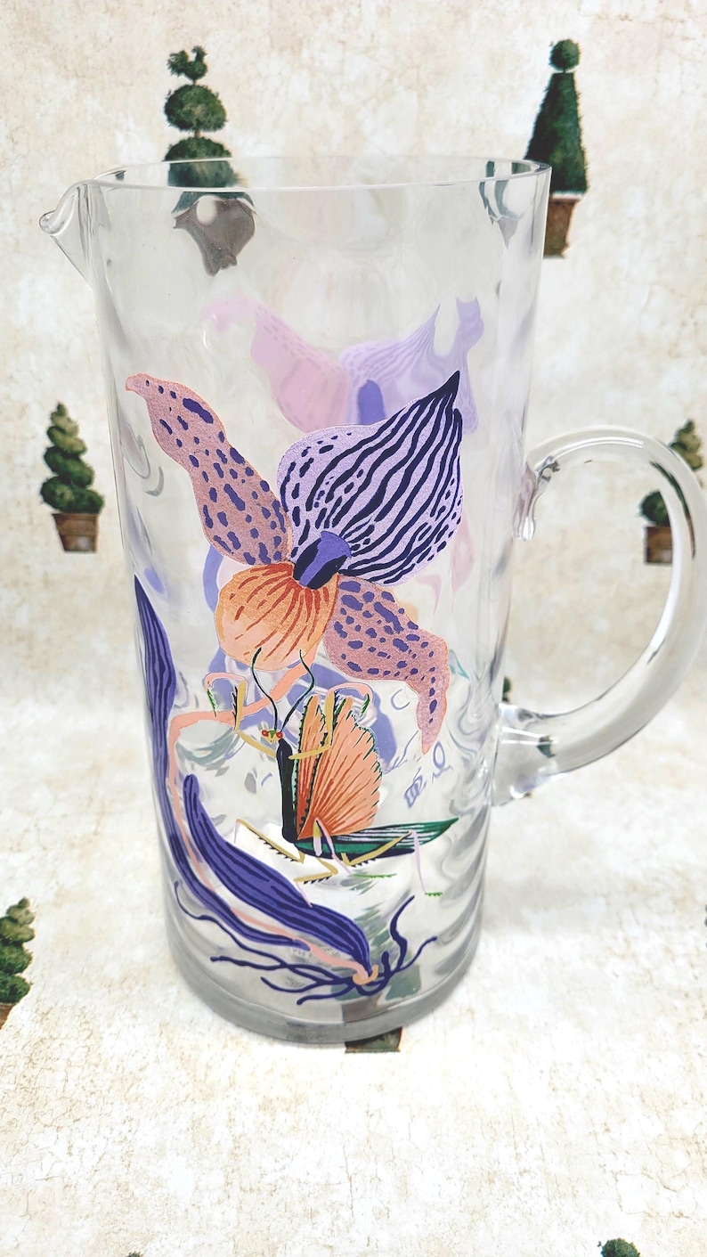 Puede incluir: Jarra de vidrio transparente con un dise&ntilde;o floral en tonos rosa, morado y naranja. La jarra tiene un asa curva y un pico vertedor. El dise&ntilde;o floral incluye una mariposa.