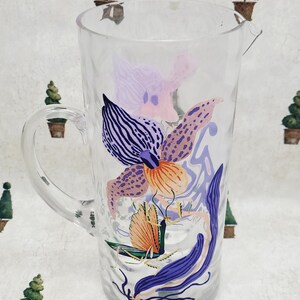 Puede incluir: Jarra de vidrio transparente con asa y boquilla, decorada con un colorido dise&ntilde;o floral. El dise&ntilde;o presenta ilustraciones de flores y hojas moradas, naranjas y azules. La jarra es adecuada para servir bebidas.