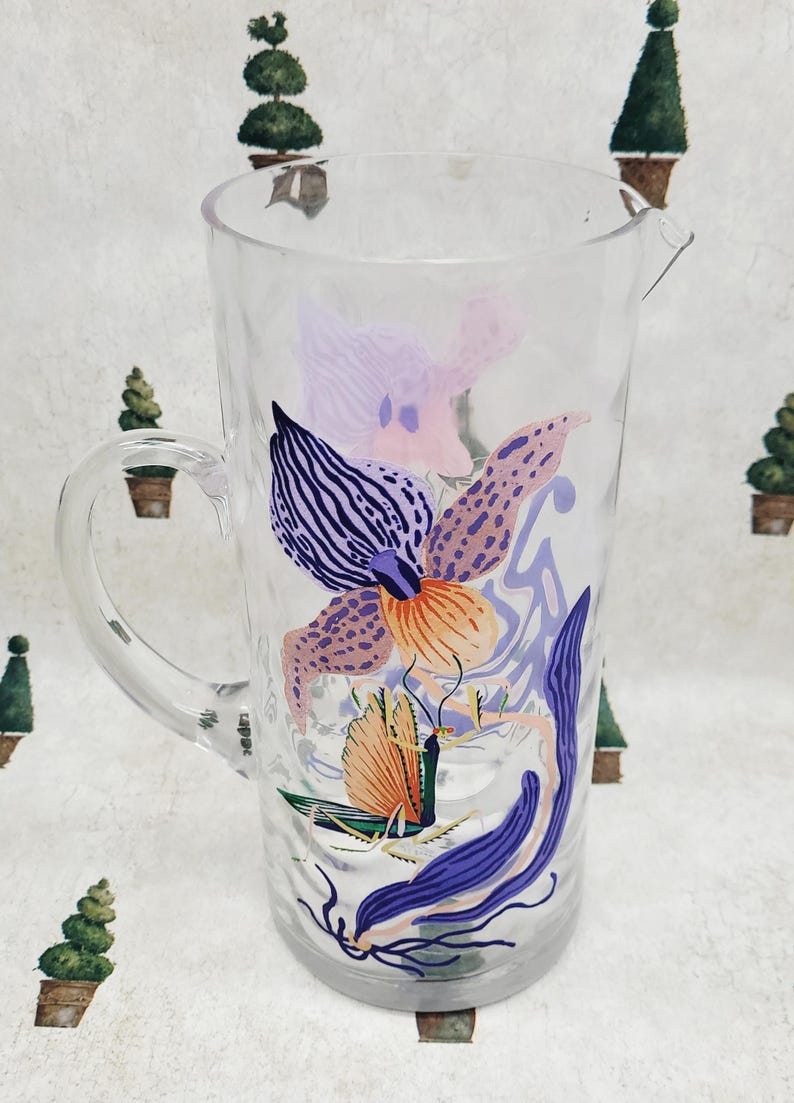 Puede incluir: Jarra de vidrio transparente con asa y boquilla, decorada con un colorido dise&ntilde;o floral. El dise&ntilde;o presenta ilustraciones de flores y hojas moradas, naranjas y azules. La jarra es adecuada para servir bebidas.