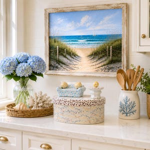 May include: A coastal kitchen counter display featuring a framed beach landscape painting, a glass vase with blue hydrangeas, a white coral piece, a woven tray, a patterned oval box, a small blue sofa figurine, and a ceramic utensil crock.