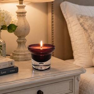 May include: A lit candle in a deep purple glass vessel with a wide, flared rim and a clear, thick base. The candle features a single burning wick, casting a warm glow from within the dark-tinted glass, which sits on a flat surface.
