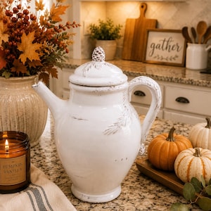 May include: A white ceramic teapot with a distressed finish, featuring a lid topped with a small pinecone finial. The teapot has a curved handle, a tapered spout, and subtle brown painted pine needle accents on the body and lid surface.