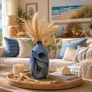 May include: A coastal-themed living room with a blue vase holding dried flowers. A wooden tray on a coffee table displays a seashell, books, and a candle. Pillows in shades of blue and white decorate the sofa, with a framed beach scene on the wall.