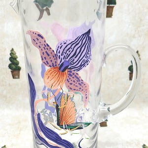 Puede incluir: Jarra de vidrio transparente con un dise&ntilde;o floral grande y colorido. La flor presenta tonos de morado, naranja y rosa. La jarra tiene un asa y un pico vertedor. El fondo tiene un patr&oacute;n repetitivo de peque&ntilde;os &aacute;rboles.