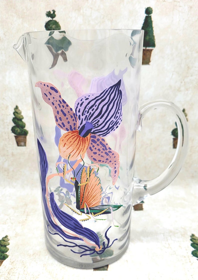 Puede incluir: Jarra de vidrio transparente con un dise&ntilde;o floral grande y colorido. La flor presenta tonos de morado, naranja y rosa. La jarra tiene un asa y un pico vertedor. El fondo tiene un patr&oacute;n repetitivo de peque&ntilde;os &aacute;rboles.