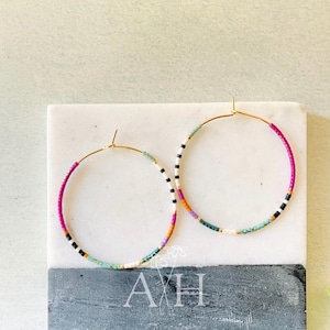 May include: A pair of gold-tone hoop earrings featuring a pattern of small, multi-colored seed beads in shades of magenta, black, white, orange, and teal. The earrings are displayed on a square block with the white text AH printed on the front.
