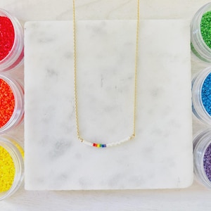 May include: A delicate gold chain necklace with a white beaded bar and a rainbow stripe. The necklace is displayed on a white marble surface, surrounded by small jars of colorful beads.