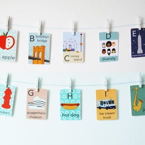 May include: A set of ten alphabet flashcards featuring illustrations of various New York City landmarks and cultural icons. Each card has a letter, a word, and a colorful illustration. The cards are hung on a string with clothespins.