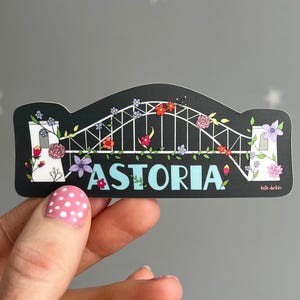 May include: A black sticker featuring a white bridge design with floral accents in red, pink, and purple. The word "ASTORIA" is in teal letters across the bottom. The sticker has a unique shape.