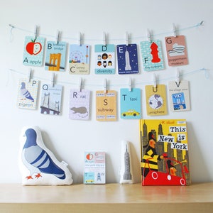 May include: A set of alphabet flashcards featuring illustrations of New York City landmarks and icons. The flashcards are hung on a string with clothespins. A stuffed pigeon and a book titled "This New is York" are on a wooden surface.
