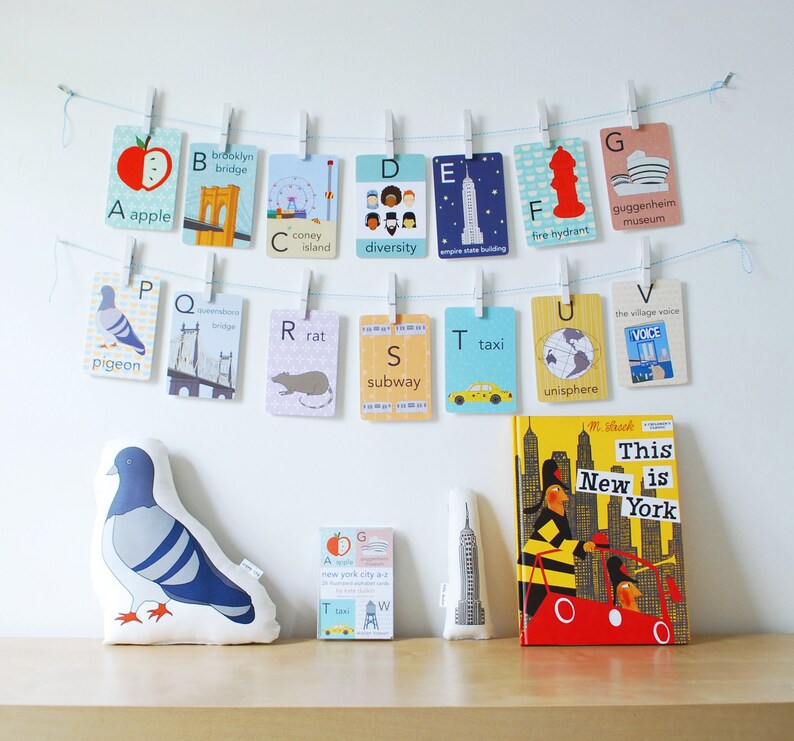May include: A set of alphabet flashcards featuring illustrations of New York City landmarks and icons. The flashcards are hung on a string with clothespins. A stuffed pigeon and a book titled "This New is York" are on a wooden surface.