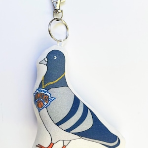 May include: A fabric pigeon keychain featuring a blue and grey bird wearing a gold chain with a New York Knicks basketball logo pendant. The plush pigeon is attached to a silver metal lobster clasp and a circular split ring for easy attachment.