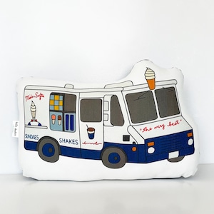 May include: A white ice cream truck pillow with blue and red details. The truck has a sign that says "Milder Softie" and offers "Sundaes" and "Shakes". The truck has a cone of ice cream on top and the text "the very best" on the front.