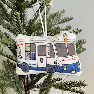 May include: A fabric Christmas ornament shaped like an ice cream truck. The ornament is white with blue and gray details, featuring text that reads "the very best", "shakes", and "sundaes". An ice cream cone is on top.