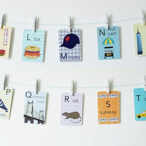 May include: A set of alphabet flashcards featuring New York City themed illustrations. Each card has a letter, a word, and a picture. For example, the card for 'K' has a picture of Katz's Delicatessen and the word 'katz's deli'.
