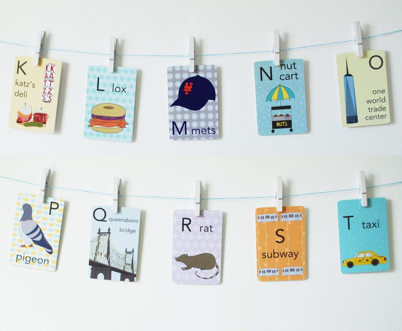 May include: A set of alphabet flashcards featuring New York City themed illustrations. Each card has a letter, a word, and a picture. For example, the card for 'K' has a picture of Katz's Delicatessen and the word 'katz's deli'.