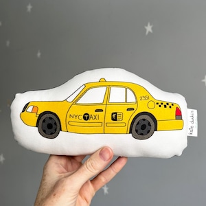 May include: A yellow toy taxi cab with the words "NYC TAXI" and the number "2351" printed on the side. The toy is made of fabric and has a white background.