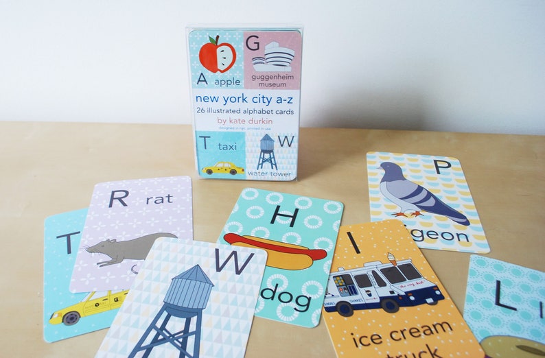 May include: A set of 26 illustrated alphabet cards featuring New York City landmarks and objects. The cards are blue, yellow, and white with illustrations of a taxi, a water tower, a hot dog, a pigeon, a rat, an apple, and an ice cream truck. The cards are titled "New York City A-Z" and are by Kate Durkin.