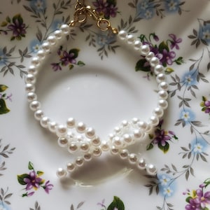 May include: A white pearl necklace with a bow detail. The necklace is on a white background with a floral pattern.