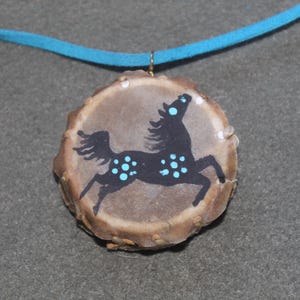 May include: A turquoise suede cord necklace with a pendant made from a round piece of antler. The antler is painted with a black silhouette of a horse with turquoise spots.