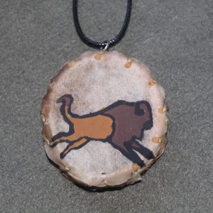 May include: A brown and black buffalo silhouette painted on a round, white leather pendant with a black cord.