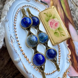 May include: Two hair clips featuring bronze-toned metal settings, each holding three round, deep blue lapis lazuli cabochons. The clips are displayed on a small, oval, white decorative tray with gold-painted edges and a rectangular floral accent.
