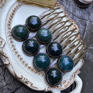 May include: A collection of vintage hair combs with dark green cabochon stones. The combs are arranged on a decorative, off-white tray with a gold trim. Other vintage items are in the background.