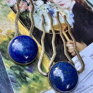 May include: Two brass hair pins with wavy stems and round, deep blue lapis lazuli stones. The pins are set against a backdrop of a painting and text that reads "BY THE SEASHORE".