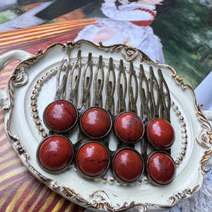 May include: A hair comb featuring eight round, polished red jasper cabochons set in a metallic bronze-toned frame. The stones display natural dark veining and are arranged in two horizontal rows of four, attached to a multi-pronged metal comb base.