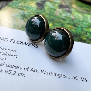 May include: A pair of dark green, round earrings with a bronze-colored metal setting. The earrings are displayed on a white surface with text, and a blurred background with a painting.
