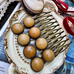 May include: An antique-style hair comb with eight amber-colored cabochons set in a bronze-toned metal. The comb is displayed on a decorative, off-white, ornate tray, alongside a cameo brooch and red ribbon. The dimensions of the image are 65 x 81.