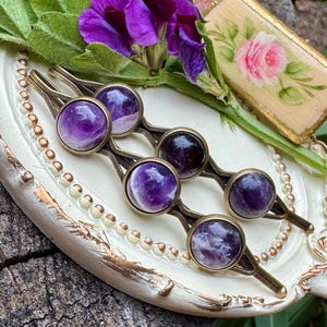 May include: Set of five antique gold hair clips with round amethyst gemstone accents. The clips are arranged on a white tray with gold accents.