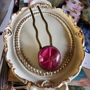 May include: A decorative hair pin with a starburst design in a deep pink hue. The pin is set in a gold-toned metal and is displayed on a cream-colored, ornate tray with gold trim. The tray has a handle.