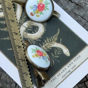 Pu&ograve; includere: Due fermagli per capelli in bronzo con accenti rotondi in porcellana bianca con motivi floreali. I motivi floreali sono in tonalit&agrave; di rosso, giallo, blu e verde. I fermagli sono su una carta con il testo "The Snake and Fern starting over".