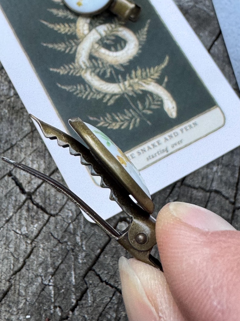 Pu&ograve; includere: Un fermaglio per capelli dorato con un design floreale bianco e giallo. Il fermaglio ha un bordo seghettato e un meccanismo a molla. Il testo "The Snake and Fern starting over" &egrave; visibile sullo sfondo.