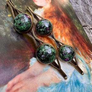 May include: Set of three hair barrettes with green and purple gemstone cabochons set in gold-toned metal. The barrettes have a geometric design and are displayed on a vintage print background. Each barrette measures approximately 2 inches in length.