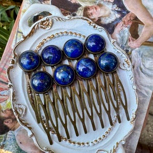 May include: A collection of eight hair combs with round, deep blue lapis lazuli stones set in brass-colored metal. The combs are arranged on a white, ornate tray with a gold trim, showcasing a vintage aesthetic.