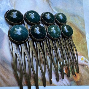 May include: A set of seven antique brass hair combs, each featuring a large, round, dark green stone cabochon. The stones have a glossy finish and are set in a brass bezel. The combs are arranged in a row.