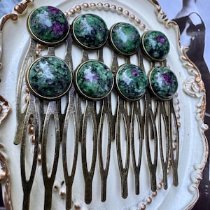May include: A set of eight hair combs with a vintage aesthetic. Each comb features a series of bronze-colored tines and is adorned with a round, polished stone. The stones display a mottled pattern of green and purple hues, set in a bronze bezel.