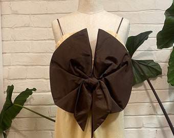 May include: A sleeveless dress with a large brown bow at the bust. The dress is a light yellow color and has a brown trim around the neckline and hem.