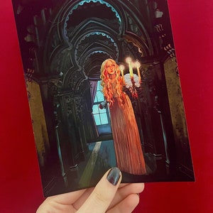 Puede incluir: Una mujer con un vestido blanco se encuentra en un pasillo oscuro con un candelabro en la mano. El pasillo está decorado con arcos ornamentados y tiene una combinación de colores azul y verde.