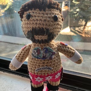 Könnte beinhalten: Handgefertigte Häkelpuppe mit braunen Haaren, Bart und schwarzen Knopfaugen. Die Puppe trägt rosa Shorts mit weißen Sternen, schwarze Stiefel mit rosa Sternen und weiße Armbänder. Auf der Shorts steht "STRAIGHT".