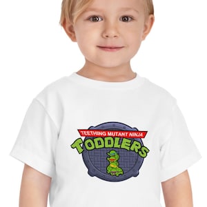 May include: White toddler t-shirt with a Teenage Mutant Ninja Turtles inspired graphic. The graphic features a green turtle with a bandana and the text "Teething Mutant Ninja Toddlers" in red and black.