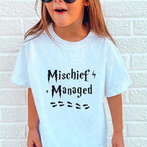 May include: A white t-shirt with black text that reads "Mischief Managed" with a lightning bolt and a star. The shirt is being worn by a child with brown hair and sunglasses.