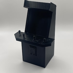 May include: A small, black arcade game cabinet replica. The miniature arcade machine features a control panel with joysticks and buttons, a screen area, and a rectangular base. The overall design is a solid, matte black.