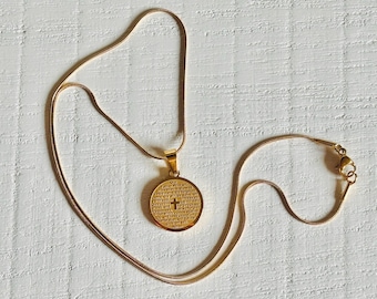 May include: Gold-colored necklace with a round pendant. The pendant features engraved text and a cross symbol. The necklace is displayed on a textured white surface. The chain is a delicate, snake chain style.