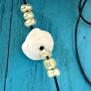 May include: A close-up of a necklace featuring a large, off-white shell pendant with a central hole, strung with gold-colored beads. The necklace is on a black cord, set against a turquoise background.