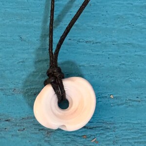 May include: A white and tan shell pendant with a dark brown cord. The shell has a circular shape with a hole in the center and a natural, organic texture. The cord is knotted above the shell.