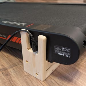 May include: Close-up of a black treadmill with a wooden support structure. The treadmill has a black belt and a digital display. The wooden support is light-colored and holds the power cord.